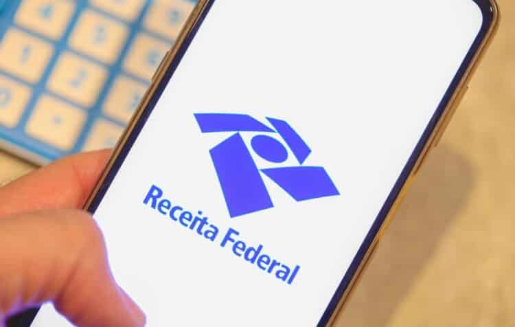 Brasilia, Federal District - Brazil. March, 25, 2023. The screen of a cell phone that a man is holding showing the Federal Revenue of Brazil logo. A calculator in the composition.
